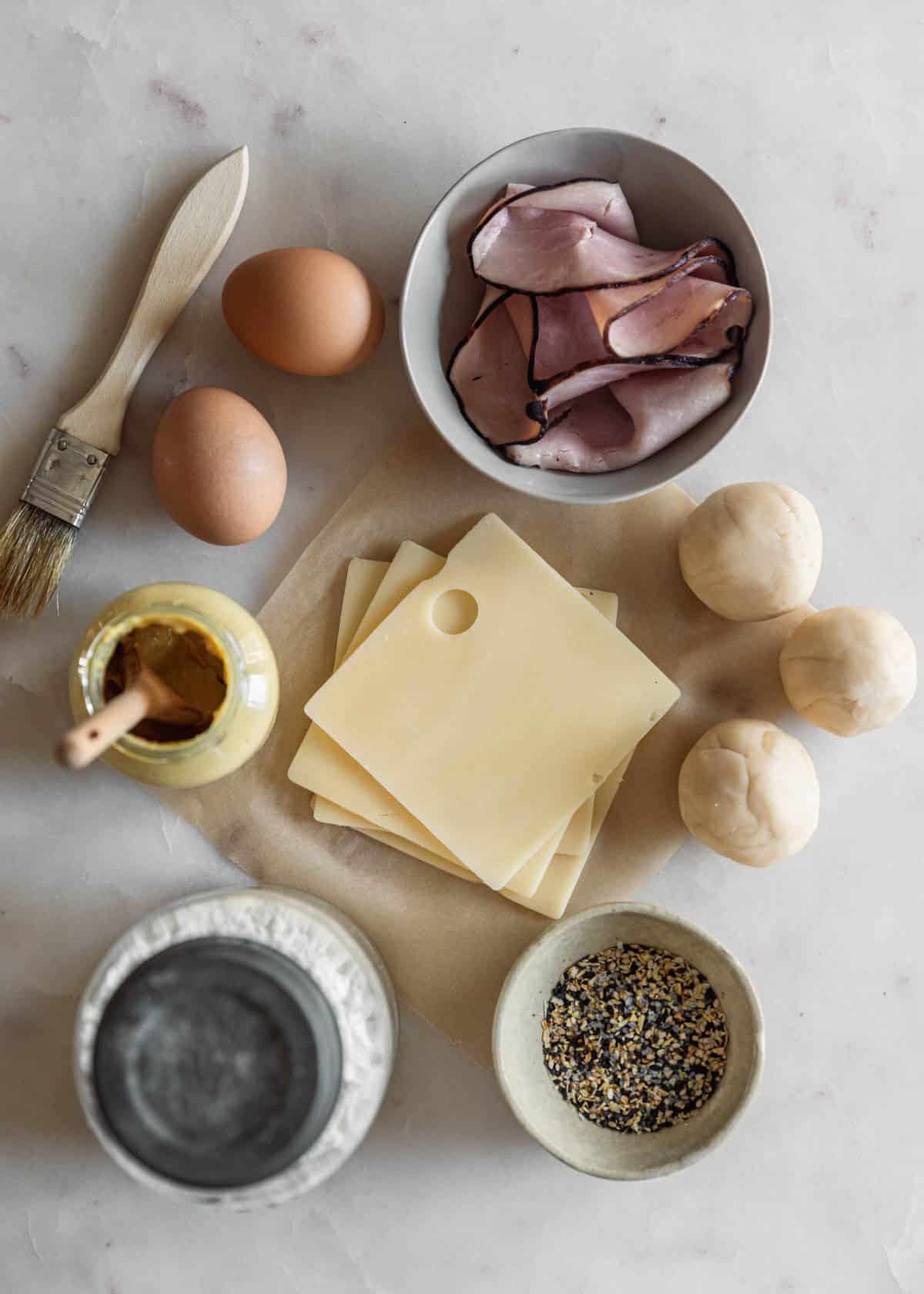 An overhead image of a square of brown parchment paper topped with Swiss cheese next to a grey bowl of ham, pie dough, eggs, a jar of Dijon, a jar of flour, and a white bowl of everything bagel seasoning on a white counter.