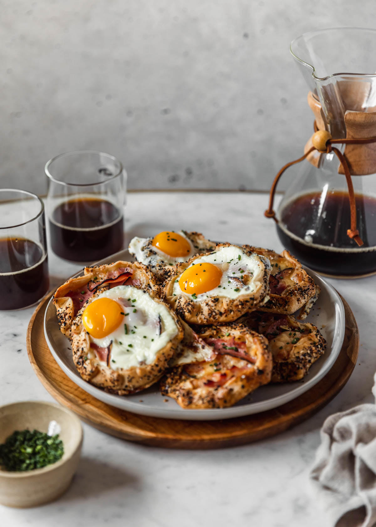 A side image of a white plate of savory croque madame galettes on a wood tray next to a Chemex, two cups of coffee, a white bowl of chives, and a beige linen on a white table with a grey speckled background.