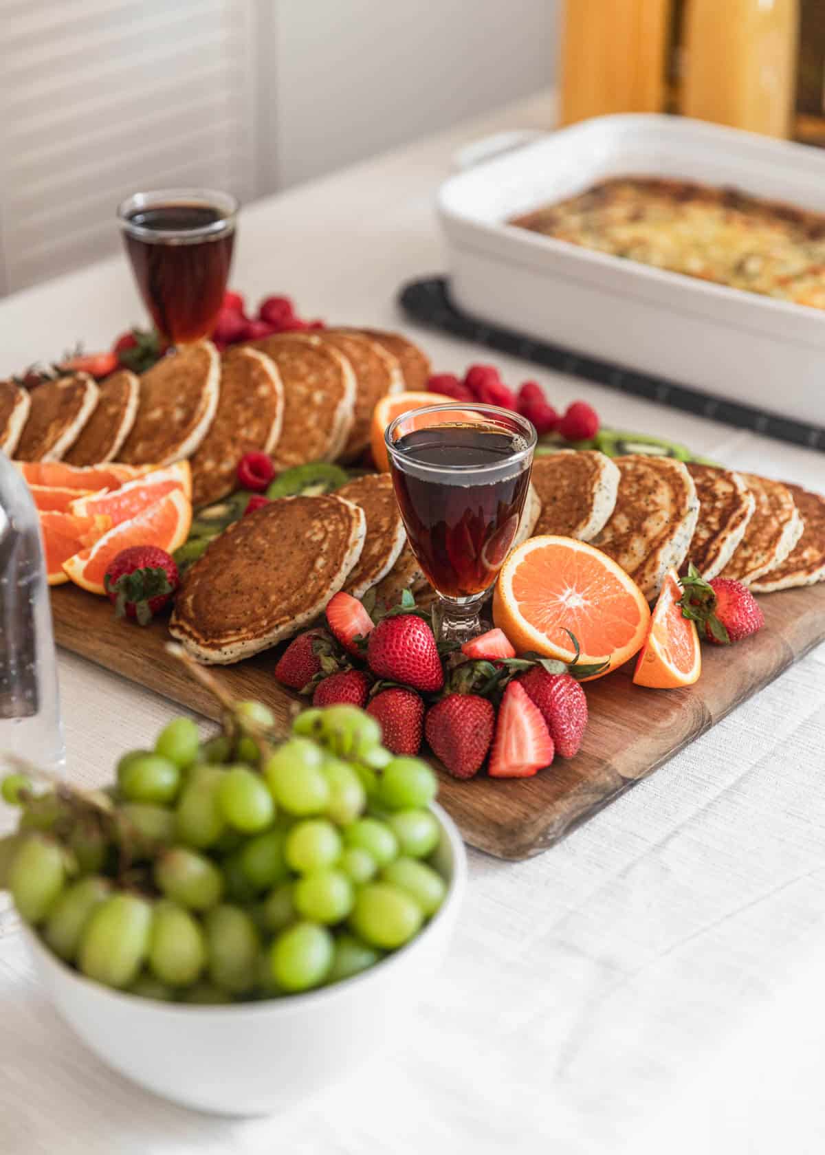 A closeup of a pancake board with fresh fruit and syrup on a white table cloth with breakfast casserole in the background.