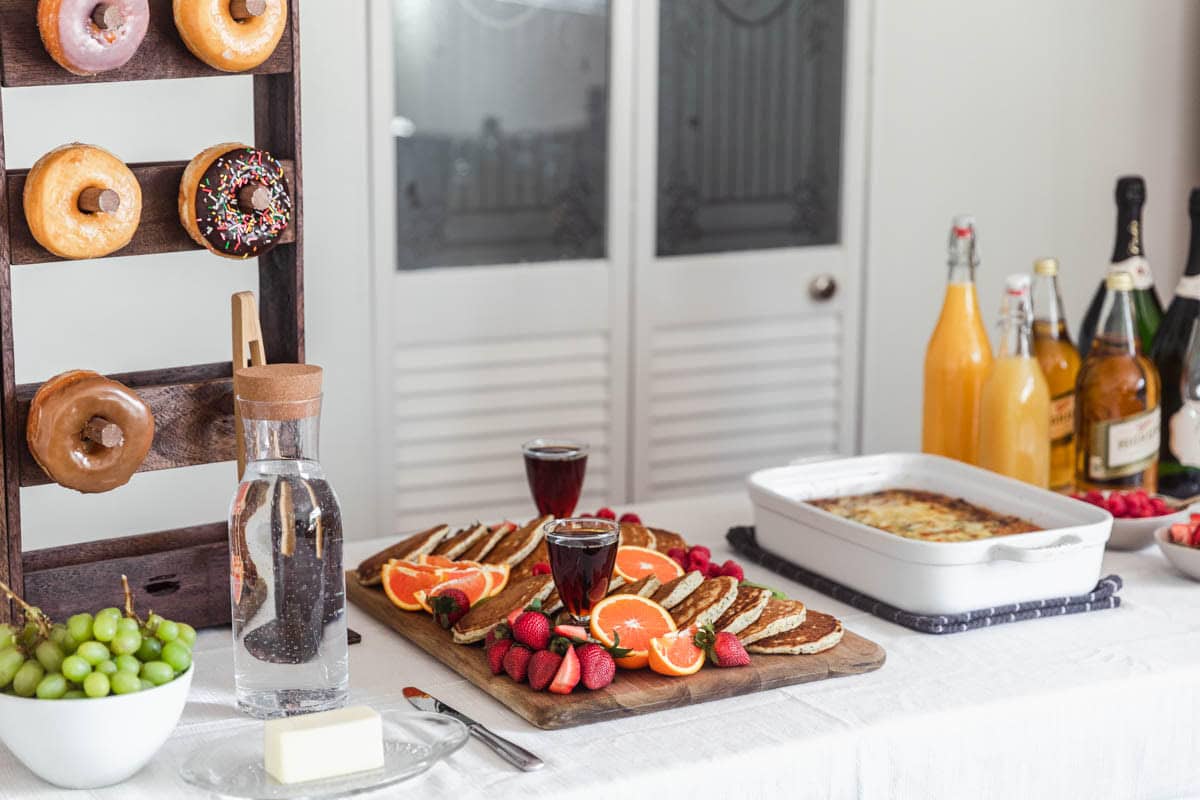 A casual brunch party set up with a pancake board, breakfast casserole, donut board, and mimosa ingredients on a white table cloth.