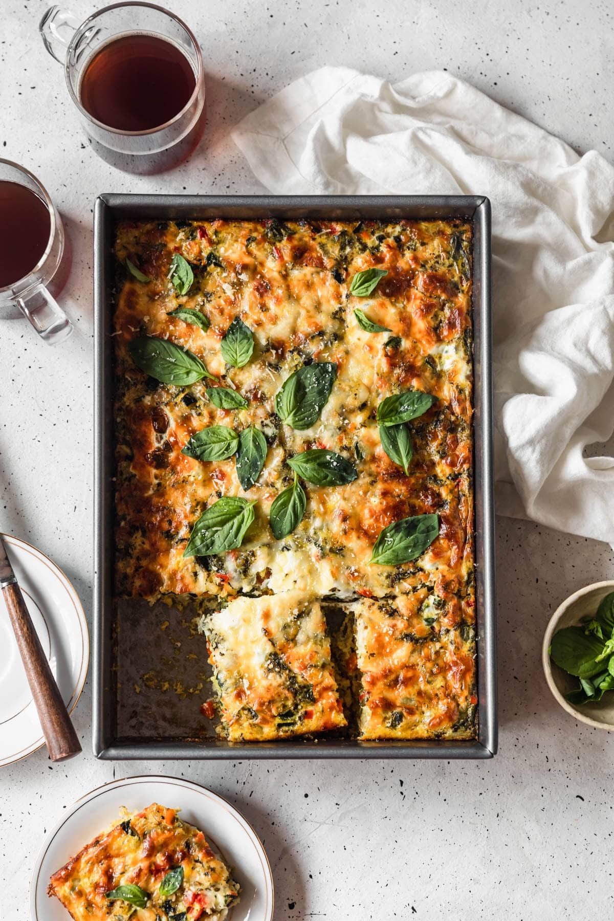 An overhead image of a metal dish with roasted veggie breakfast casserole on a white table next to a white linen, plate of casserole, and cups of coffee.