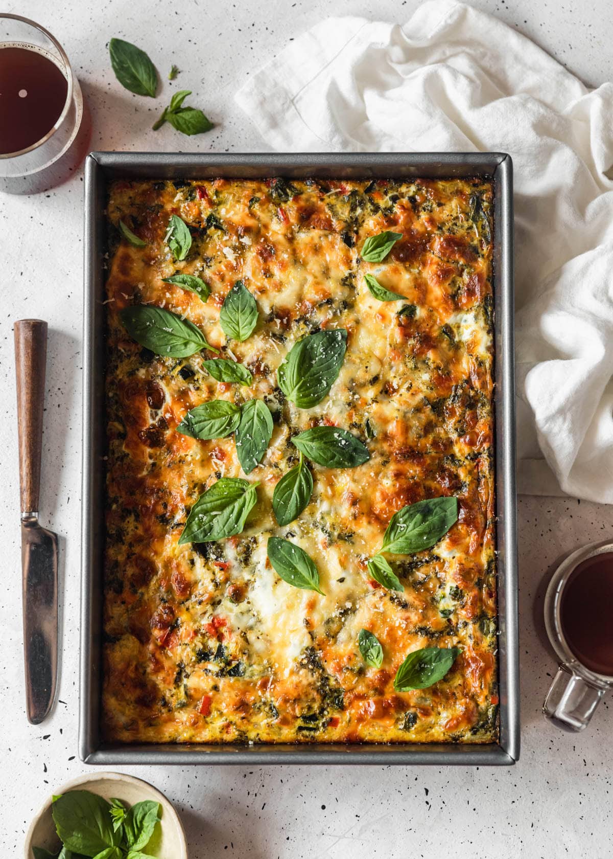 An overhead image of a roasted veggie breakfast casserole with basil on a white speckled counter next to a white bowl of basil, cups of coffee, and white napkin.