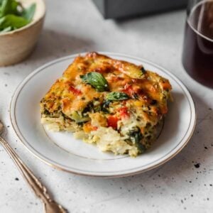 A slice of roasted veggie breakfast casserole on a white plate next to a white bowl of basil on a grey table.