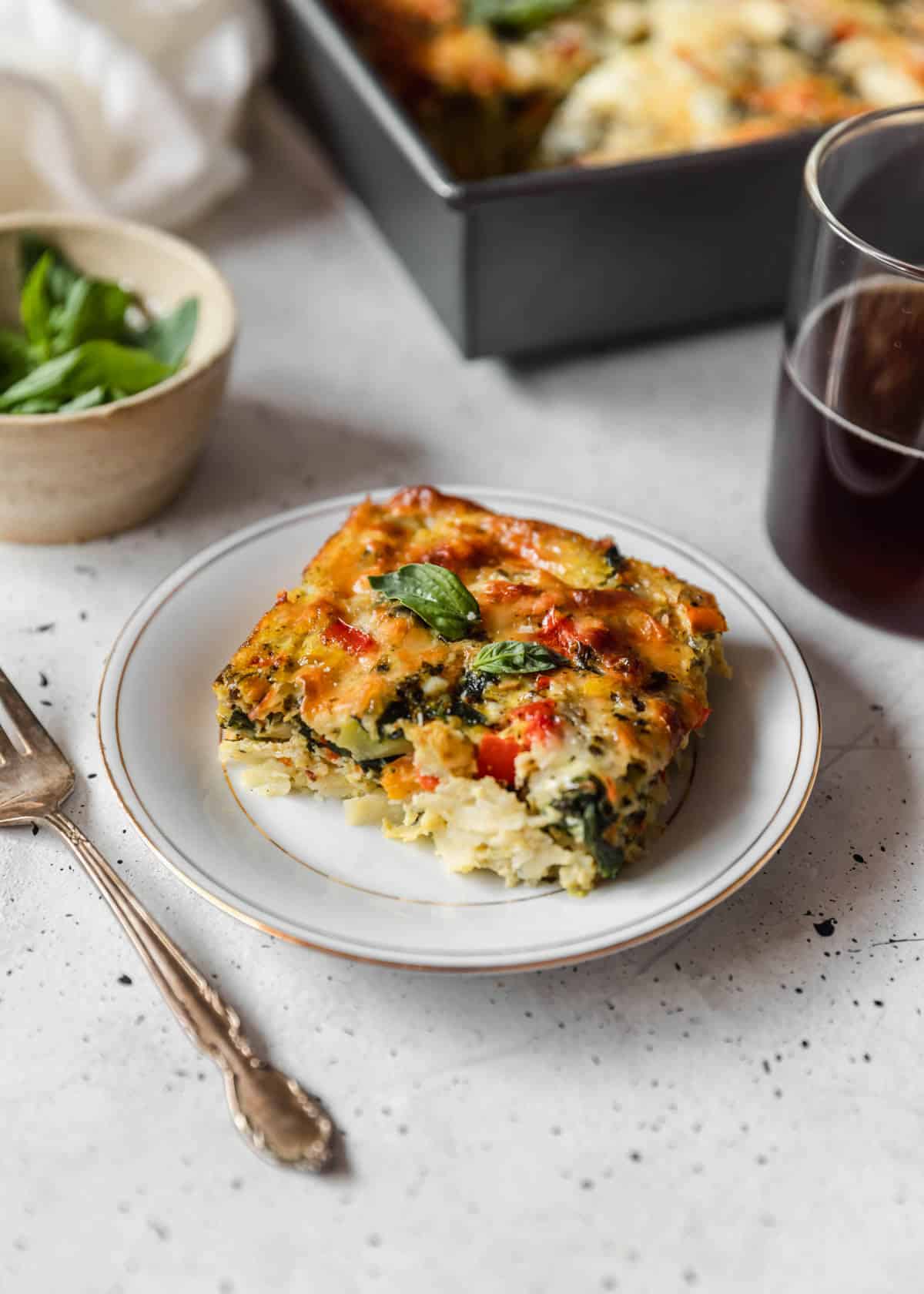 A closeup of a slice of roasted veggie breakfast casserole on a white plate next to a cup of coffee, pan, and white bowl of basil on a grey counter.