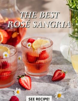A glass of rose sangria on a beige counter next to a pitcher and vase of chamomile flowers.