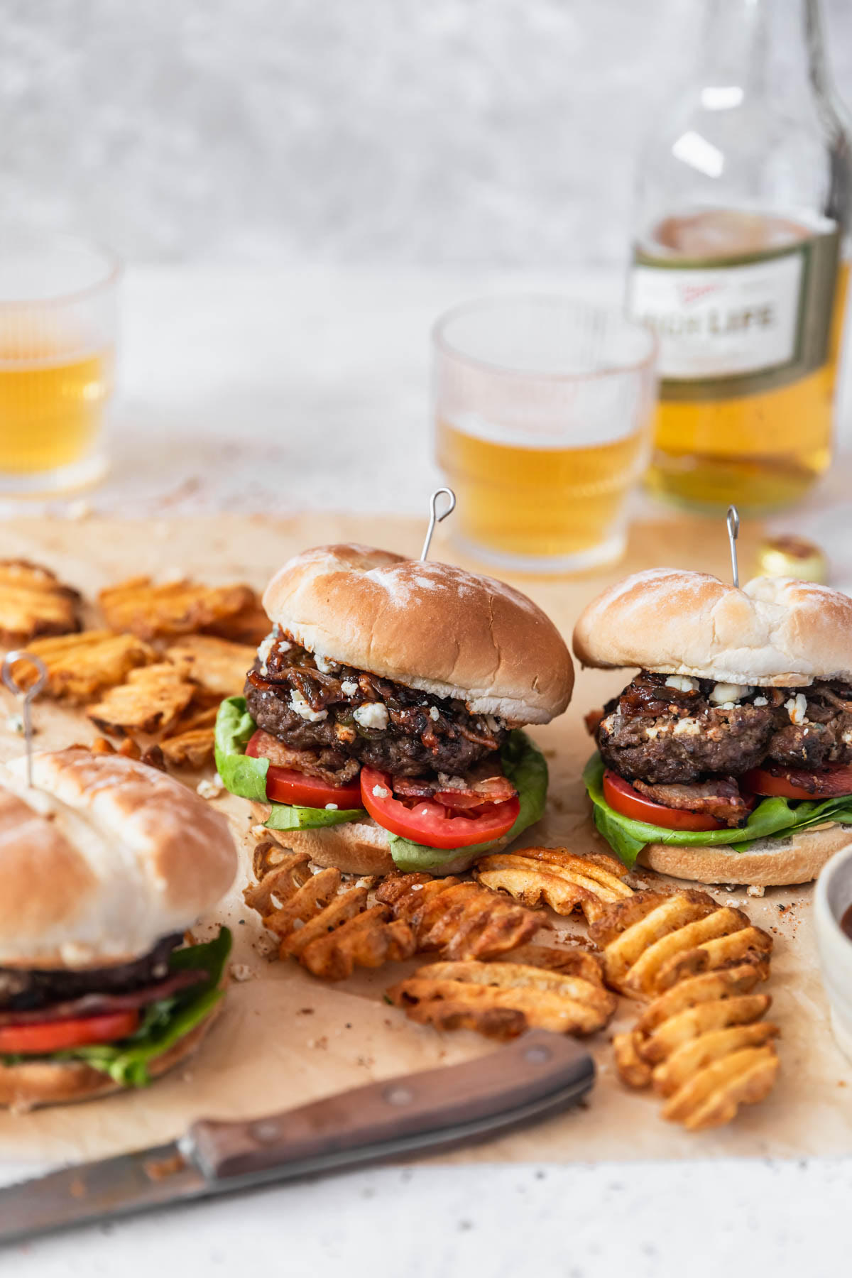 Three blue cheese burgers on brown paper next to a wood knife, waffle fries, glasses of beer, and a bottle of beer with a light grey background.