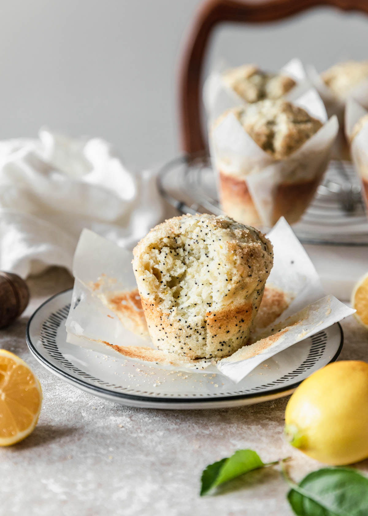 A close shot of a ricotta lemon poppy seed muffin with a bite taken out of it on a white plate with more muffins and lemons in the background on a beige table.