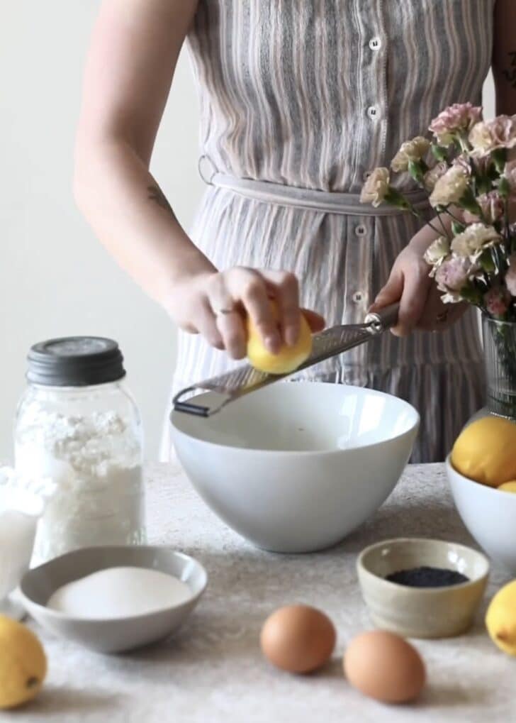 A woman in a grey and pink dress zesting lemons into a white bowl on a tan counter next to white and brown bowls of baking ingredients.