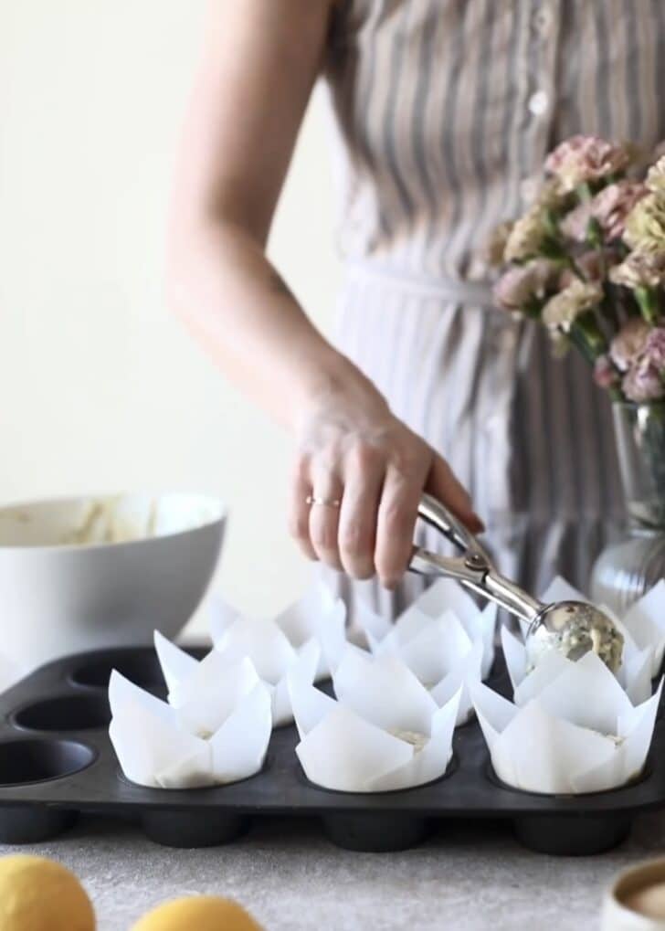 A woman in a grey and pink dress scooping ricotta lemon poppy seed muffins batter into liners on a beige counter next to pink flowers and lemons.