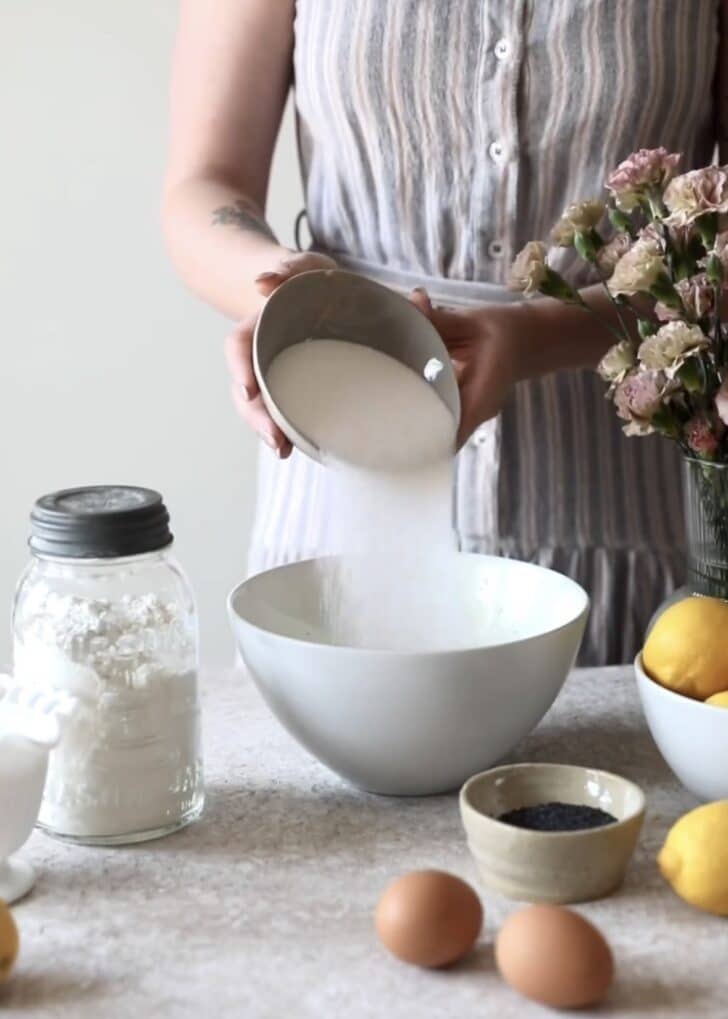 A woman in a grey striped dress pouring sugar into a white bowl on a beige counter next to baking ingredients.