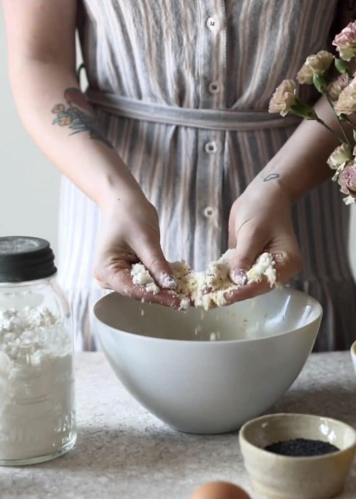 A woman in a grey and pink striped dress rubbing sugar and lemon zest in a white bowl on a beige counter next to a jar of flour.