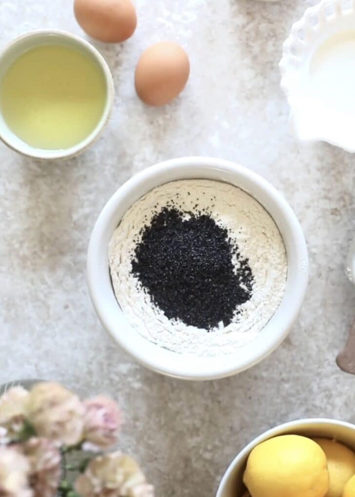 A white bowl of flour and poppy seeds on a beige counter next to pink flowers, a white bowl of lemons, and a white bowl of oil.