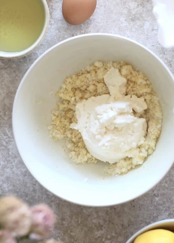 A white bowl of lemon sugar and ricotta on a beige counter next to a white bowl of oil and eggs.