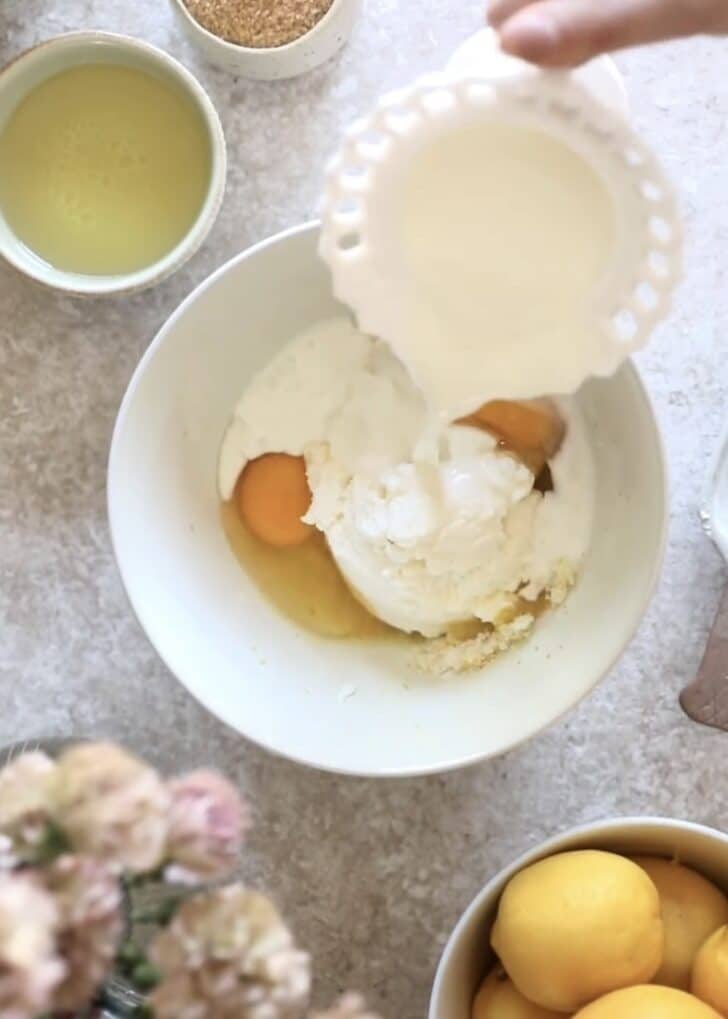 A hand pouring milk into a white bowl with muffin ingredients on a beige counter next to a bowl of lemons and pink flowers.