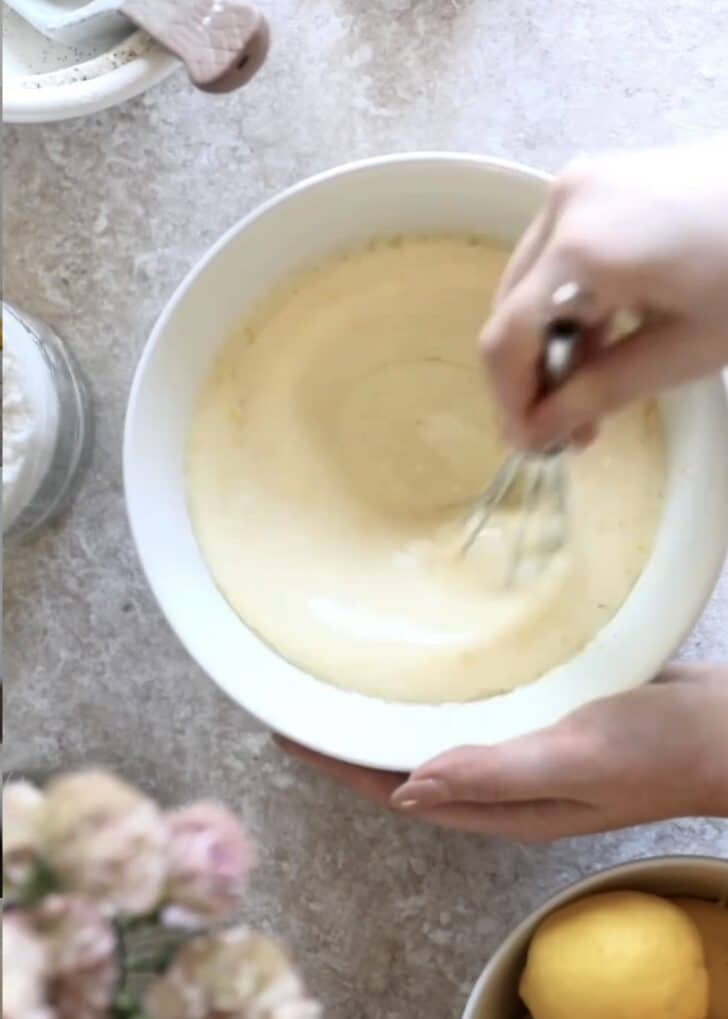 Hands whisking eggs, milk, and sugar in a white bowl on a beige counter next to lemons and pink flowers.