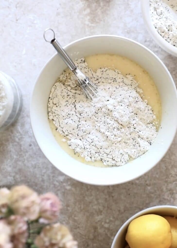 A white bowl with whisked sugar, eggs, and milk topped with flour on a beige counter next to lemons and pink flowers.