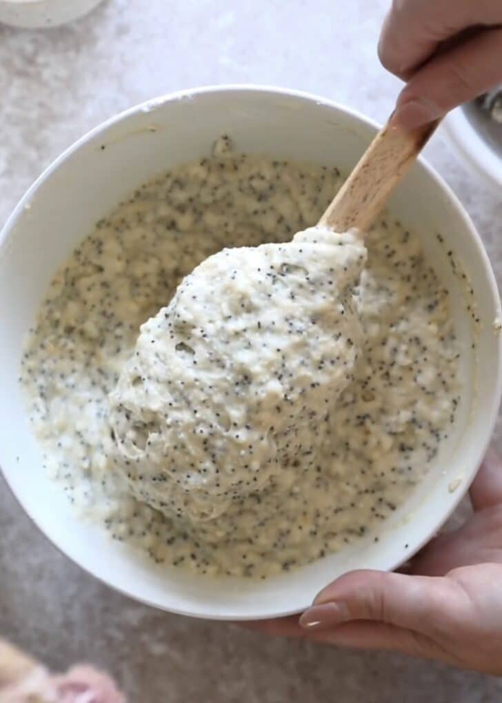 A hand using a rubber spatula to mix ricotta lemon poppy seed muffins batter in a white bowl.