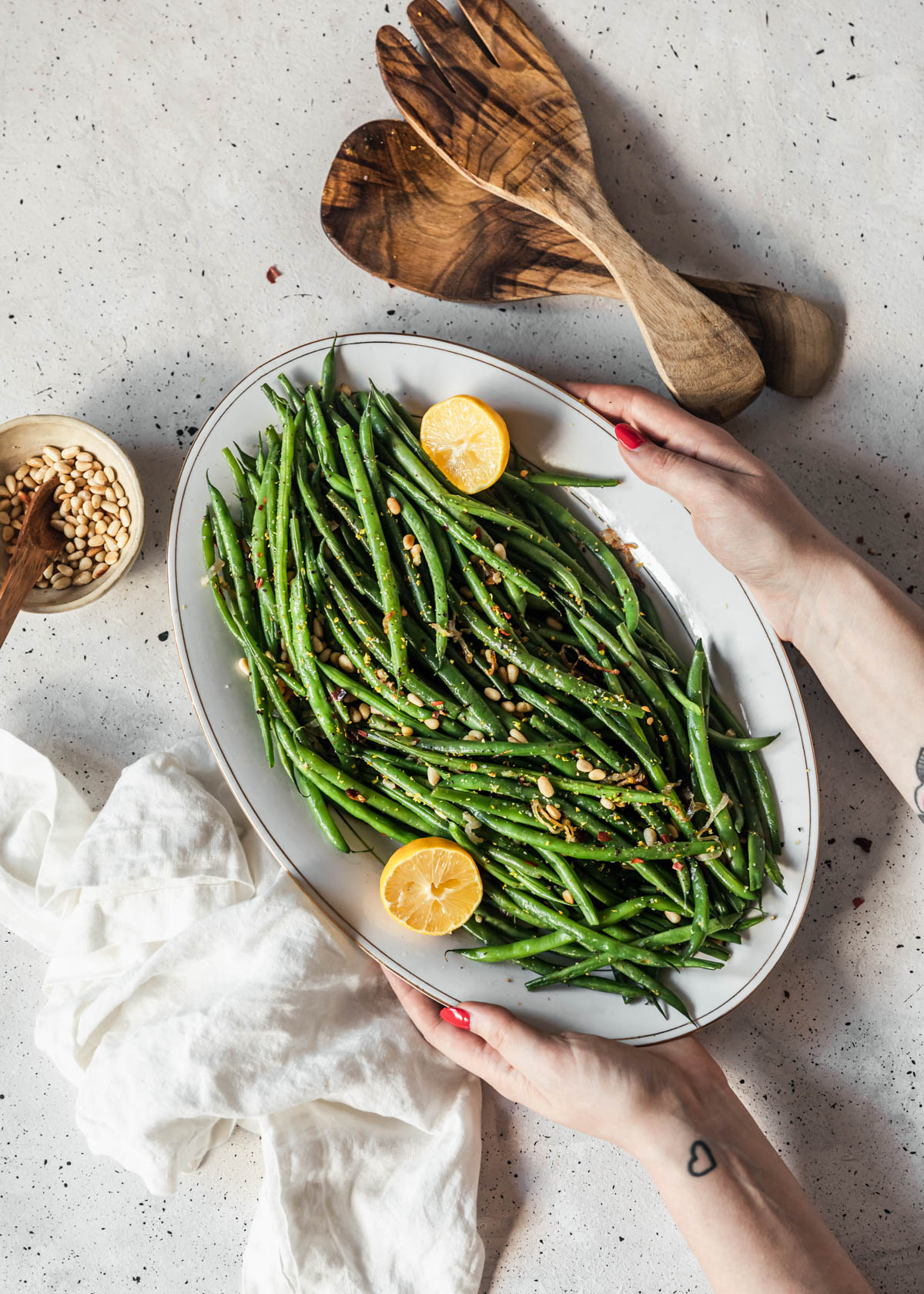 A woman's hands holding a white platter with lemony green beans over a white counter next to wood salad tossers and a bowl of pine nuts.