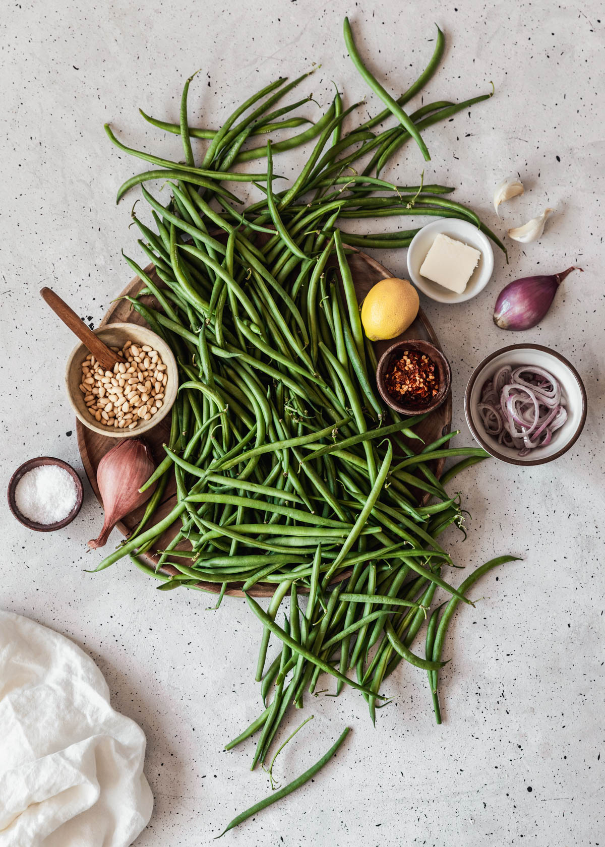 A wood board topped with string beans, a bowl of pine nuts, shallots, lemon, and garlic on a light grey table.