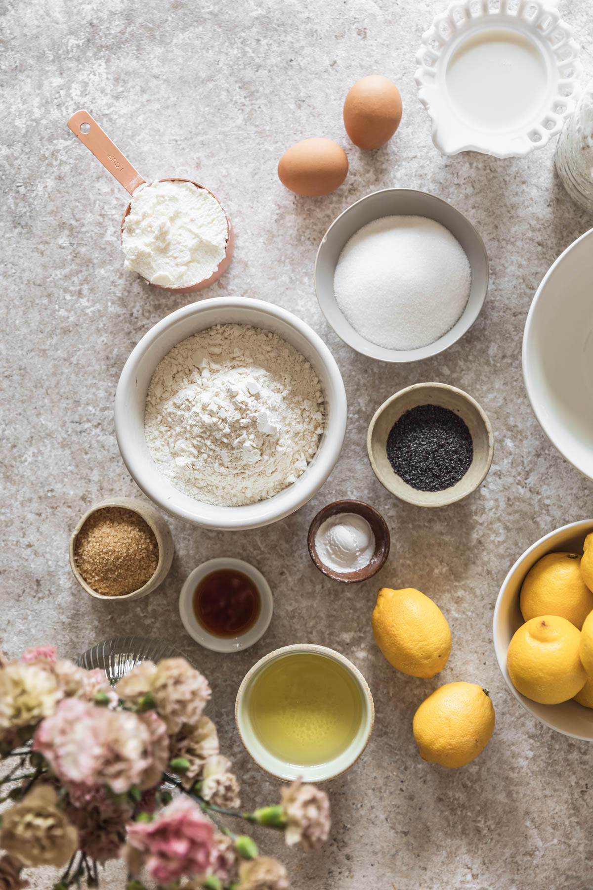 Various white and grey bowls with baking ingredients on a beige table next to a bouquet of pink flowers and white bowl of lemons.