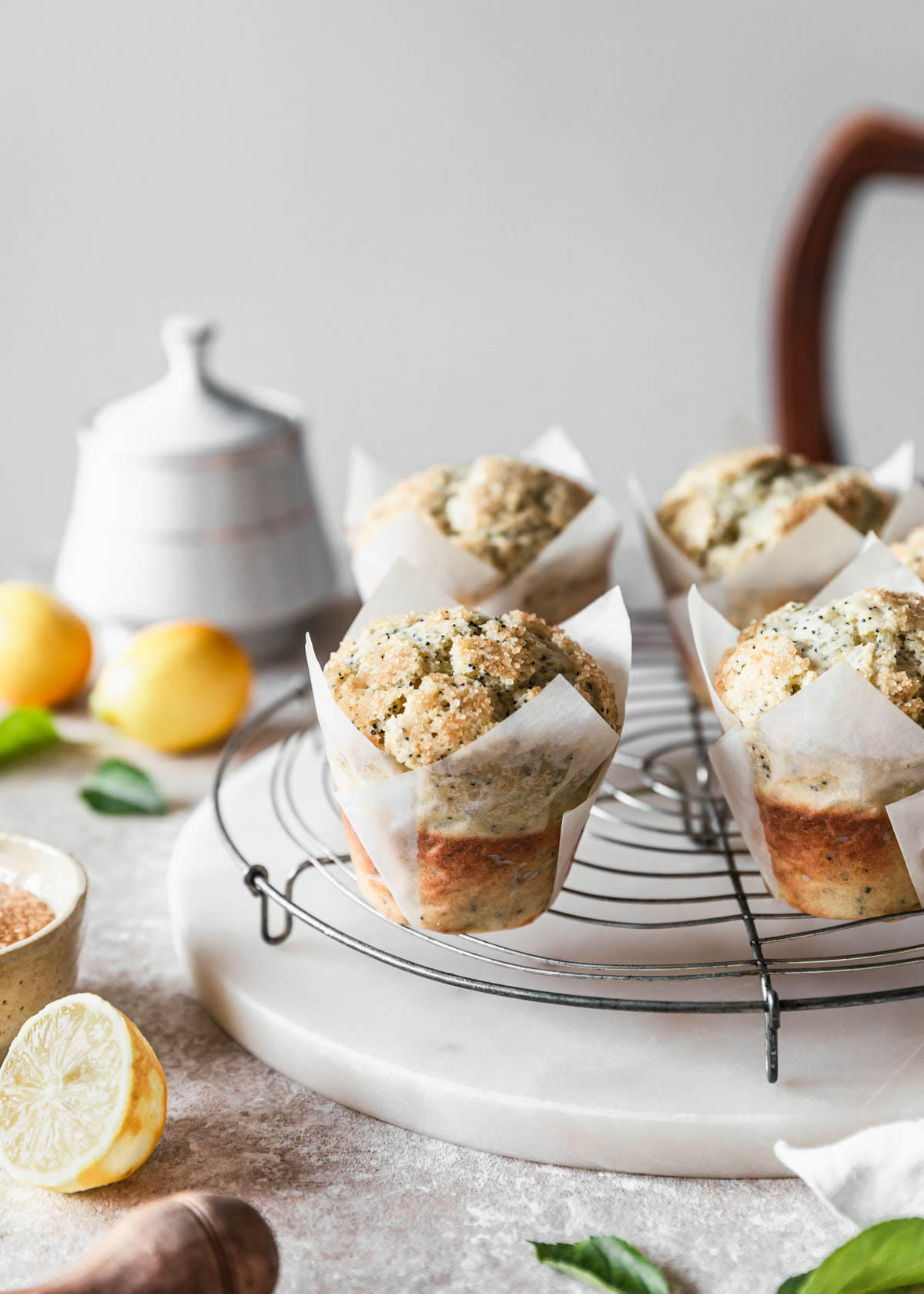 Ricotta lemon poppy seed muffins on a circular cooling rack next to lemons and a white sugar bowl on a beige speckled counter.