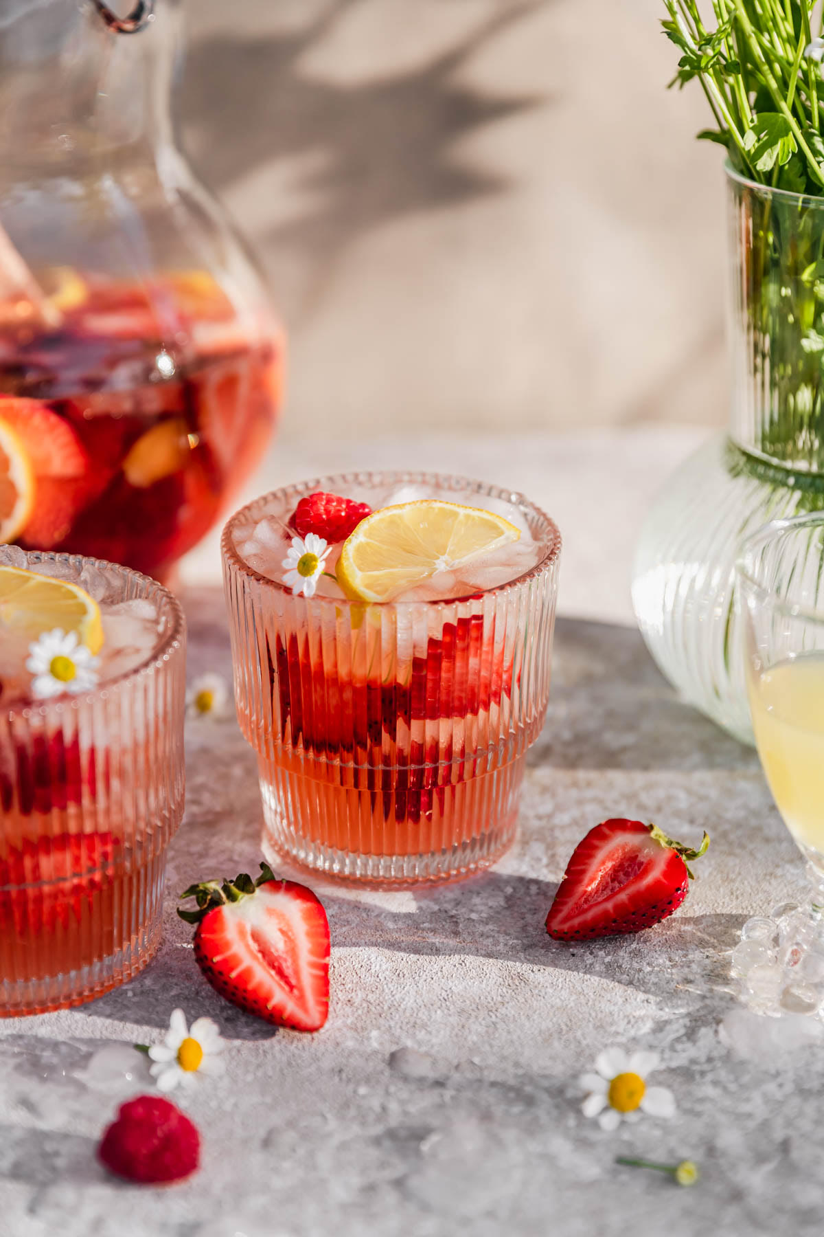 Two glasses and a pitcher with rosé sangria with berries and lemon on a tan counter next to strawberries and a vase of daisies.