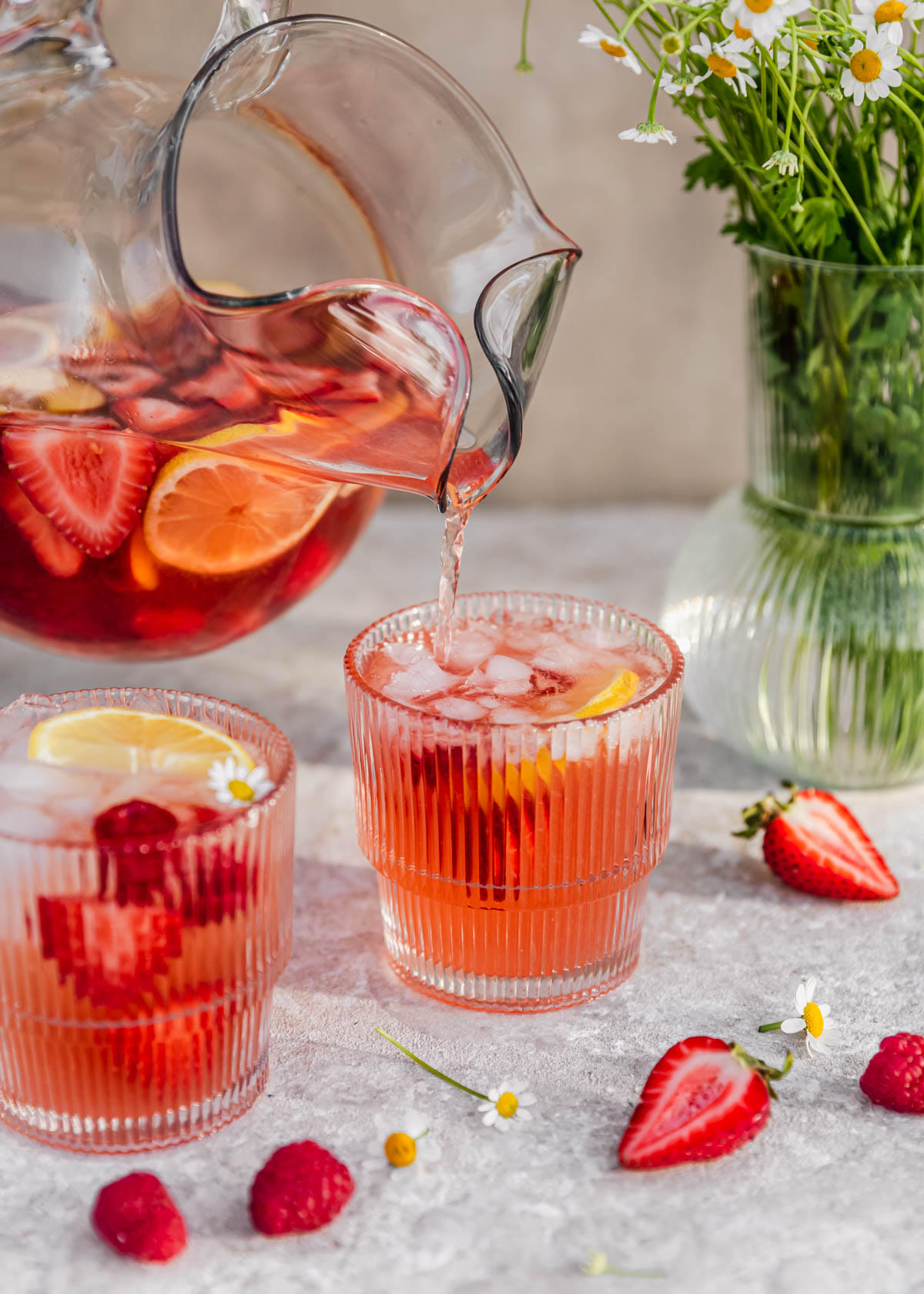 Rosé sangria being poured from a pitcher into a glass next to another glass of sangria and a vase of white flowers on a beige background.