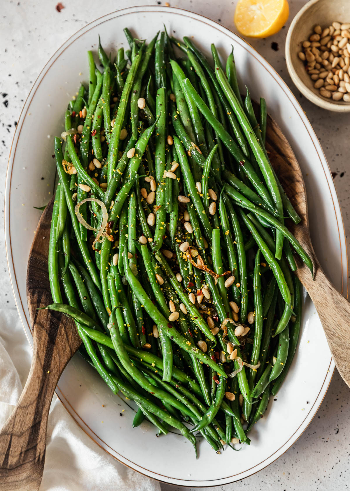 Lemony green beans with pine nuts and garlic on a white oval tray with wood salad tongs placed on a white speckled counter.