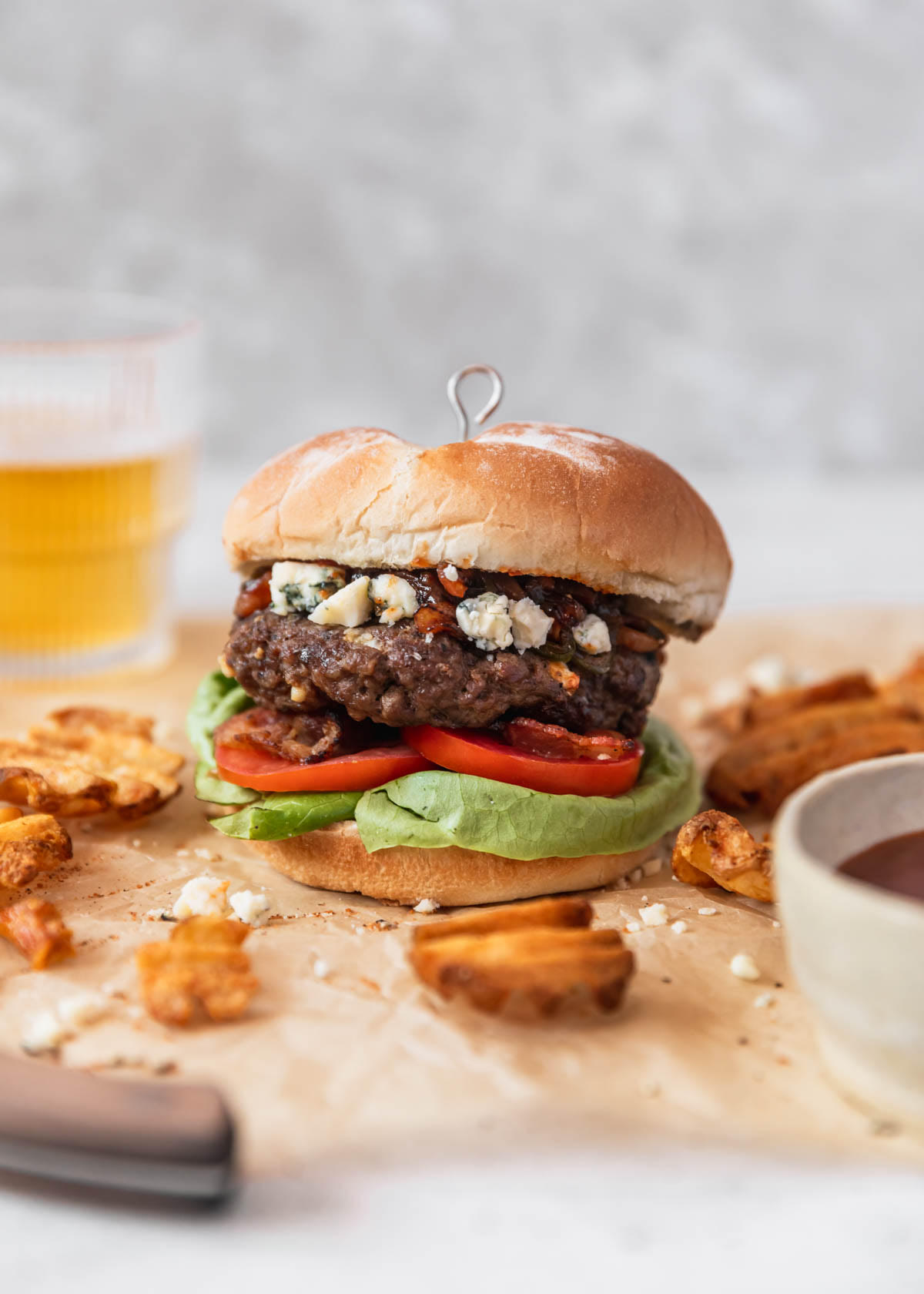 A blue cheese burger with bacon, tomato, and lettuce on tan parchment paper next to waffle fries and a glass of beer in front of a grey background.