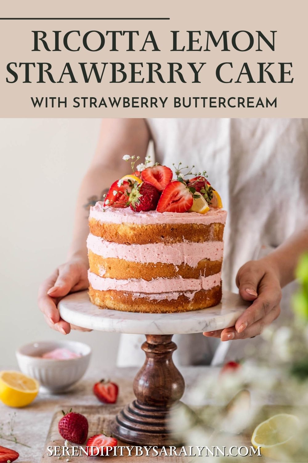 A woman with a beige apron holding a marble cake stand with ricotta strawberry lemon cake on a beige table next to a bouquet of white flowers.