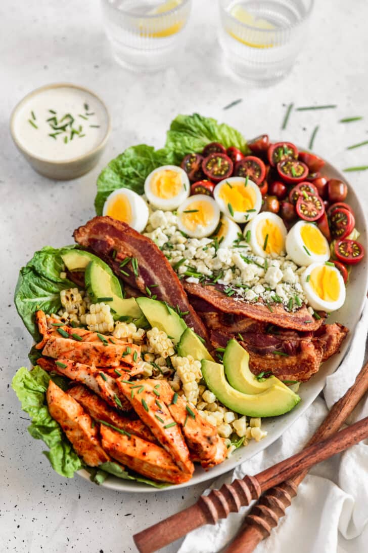 A salad with saucy chicken, corn, avocado, bacon, hard boiled eggs, tomatoes, and blue cheese on a grey counter next to wood salad tossers.