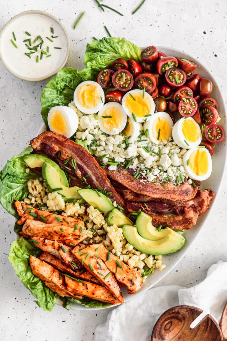 An overhead photo of a buffalo chicken Cobb salad on a white oval dish next to a white bowl of ranch on a grey speckled table.