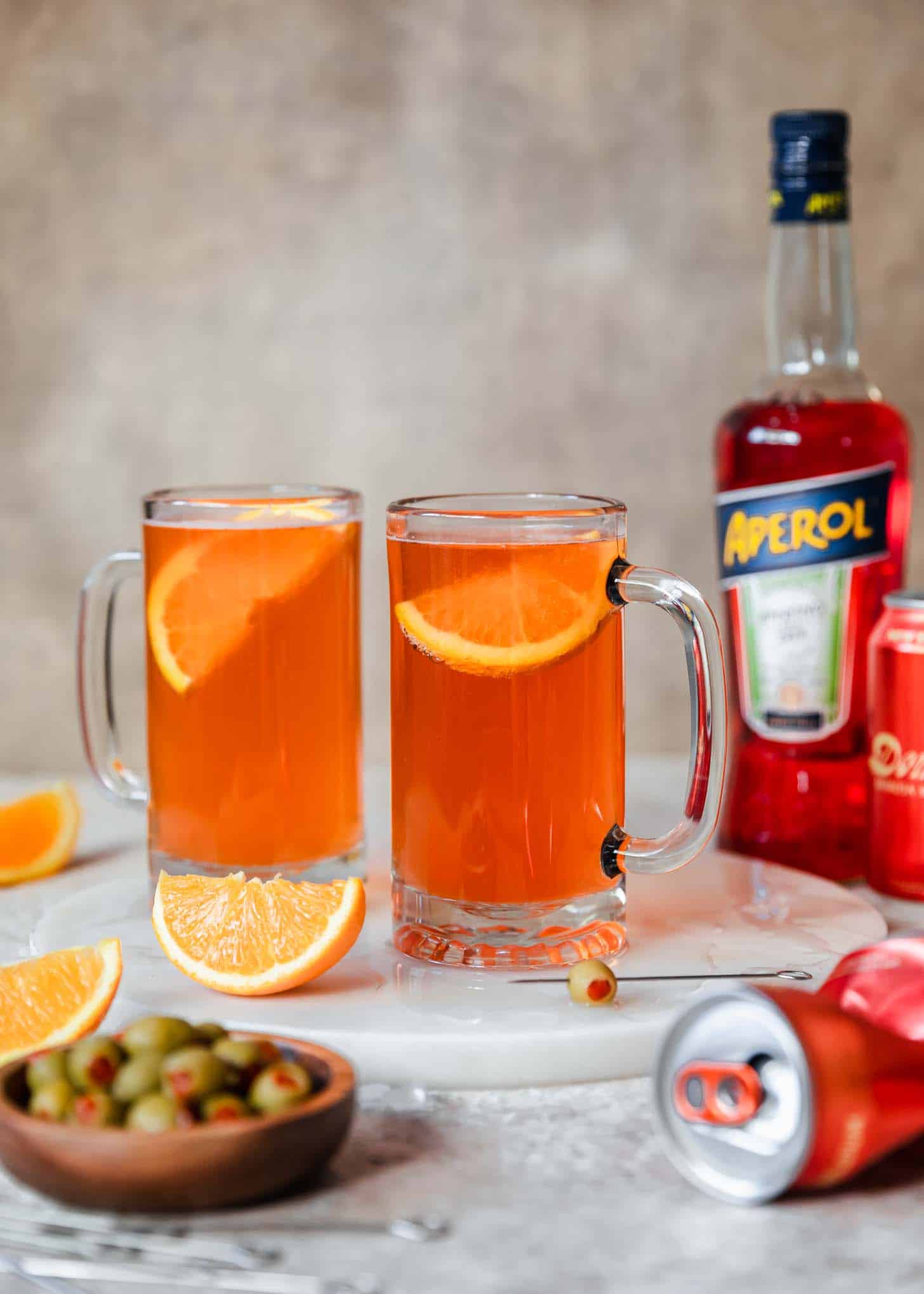 Two sour spaghett cocktails on a marble board next to a beer can, orange slices, bottle of Aperol, and wood bowl of olives with a tan background.