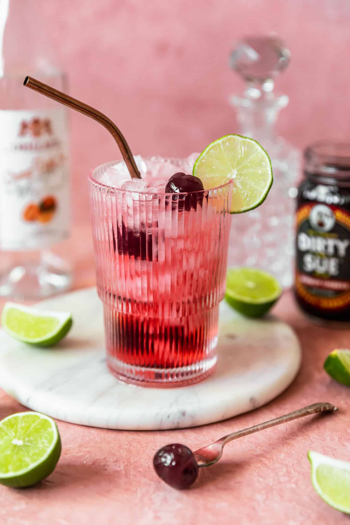 An orange dirty Shirley on a marble tray next to alcohol bottles, lime wedges, and cherries with a pink background.