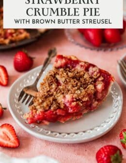 A closeup of brown butter strawberry crumble pie on a white plate next to strawberries with a pink background.