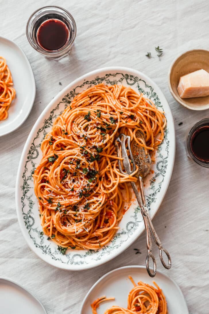 Spaghetti with roasted tomato sauce on a white oval tray next to white plates and glasses of wine on a beige linen.