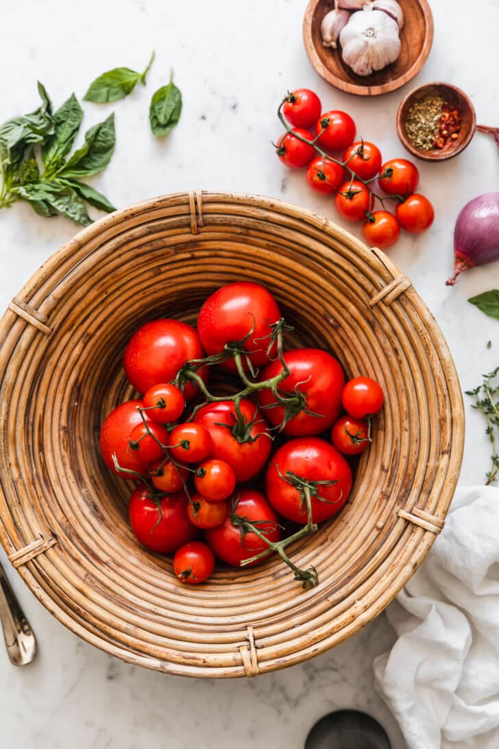 A basket of tomatoes on a white marble counter next to herbs, onions, and garlic.