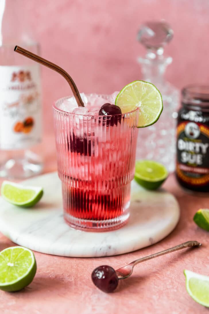 An orange dirty Shirley cocktail on a marble plate next to a jar of cherries, limes, and alcohol bottles with a pink background.