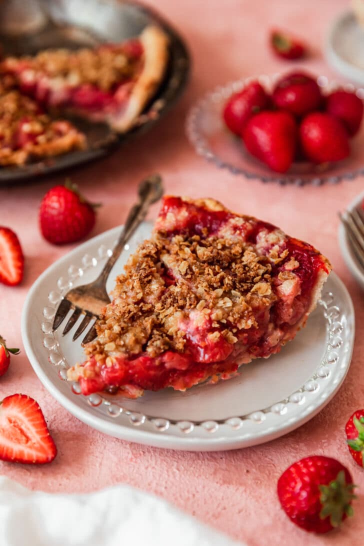 A closeup of a slice of brown butter strawberry crumble pie on a white plate next to strawberries and a full pie with a pink background.