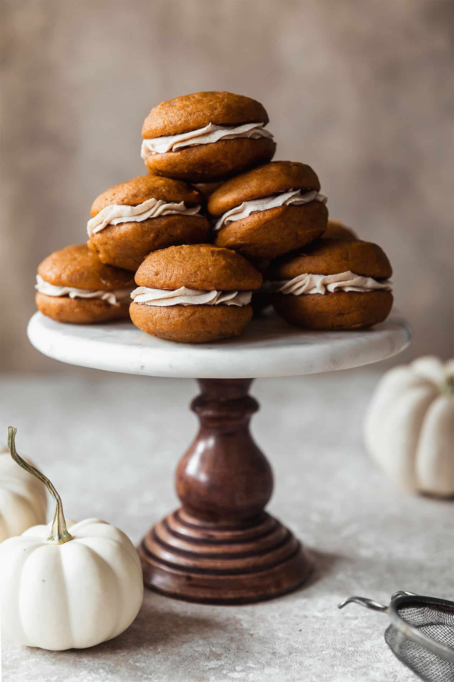 Pumpkin whoopie pies stacked on a marble and wood cake stand next to white pumpkins on a brown background.