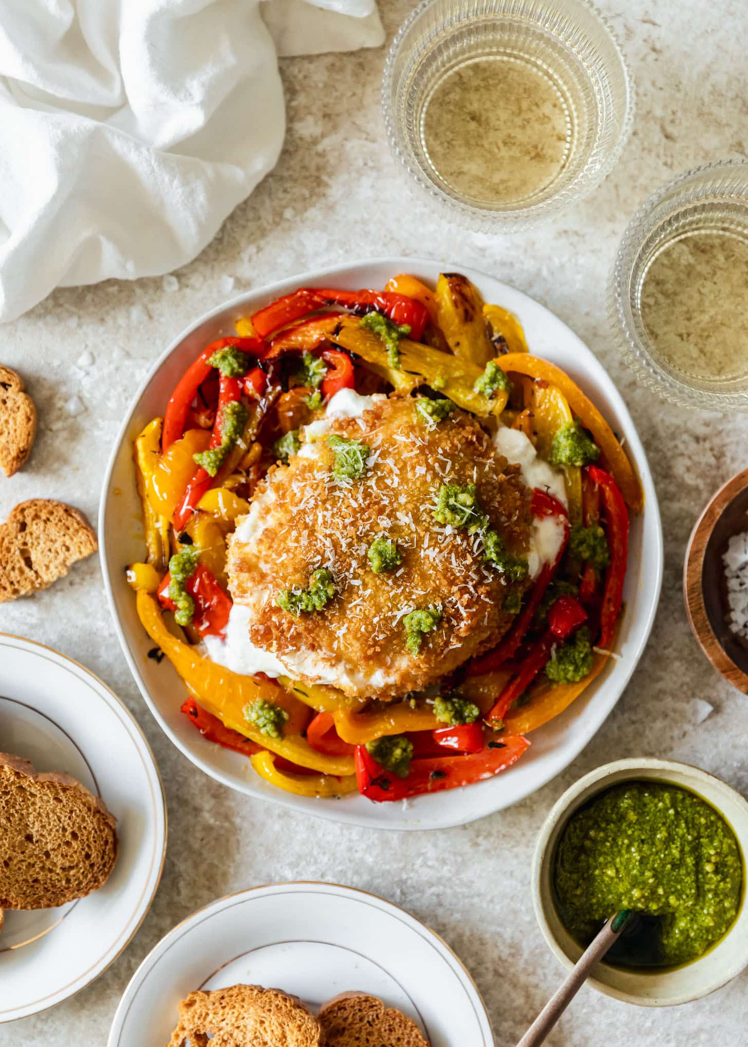 Fried burrata and peppers on a white plate next to baguette slices, white wine, pesto, and a white linen on a tan counter.