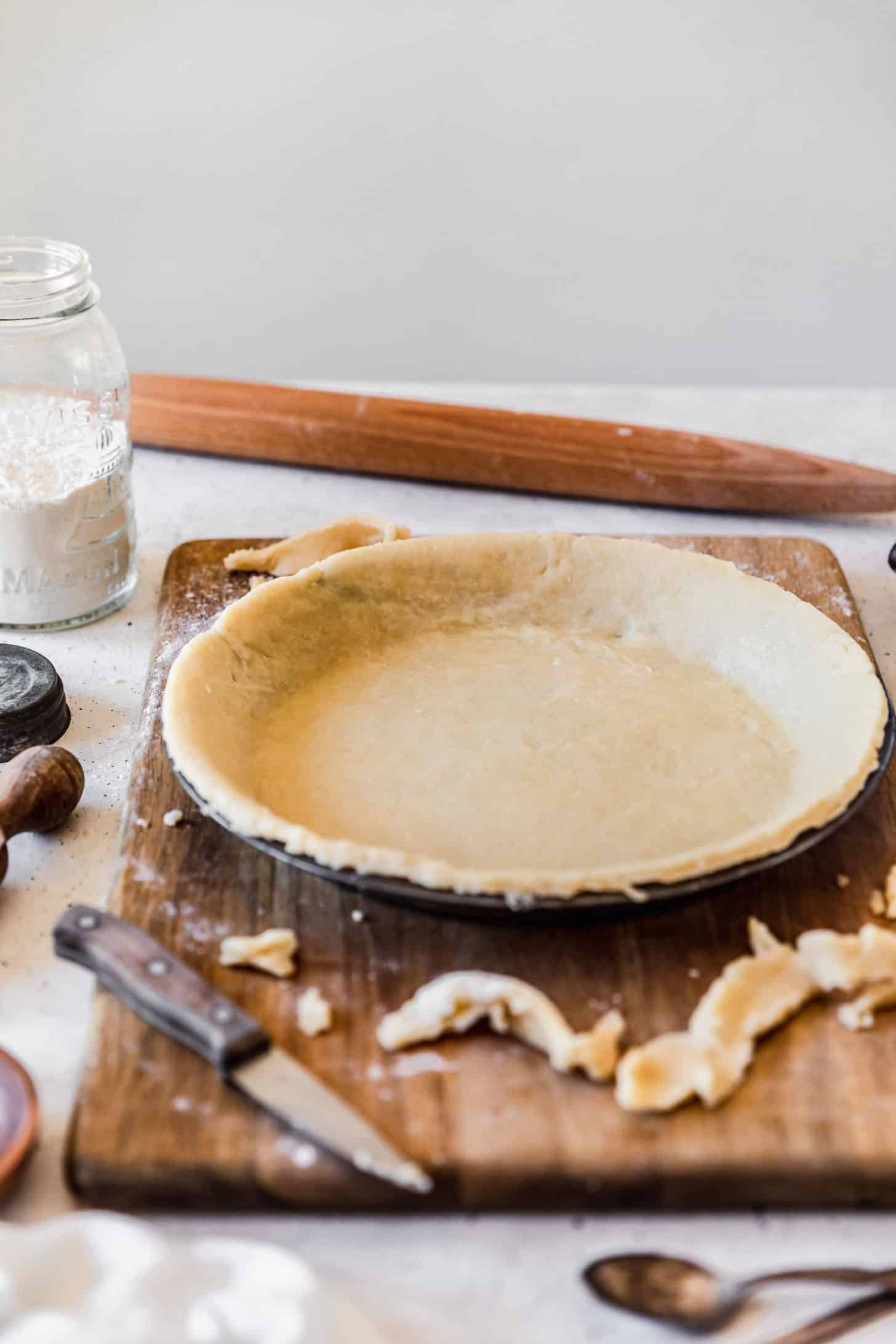 The best pie crust in a metal pie plate on a wood board next to pie tools and ingredients in front of a white wall.