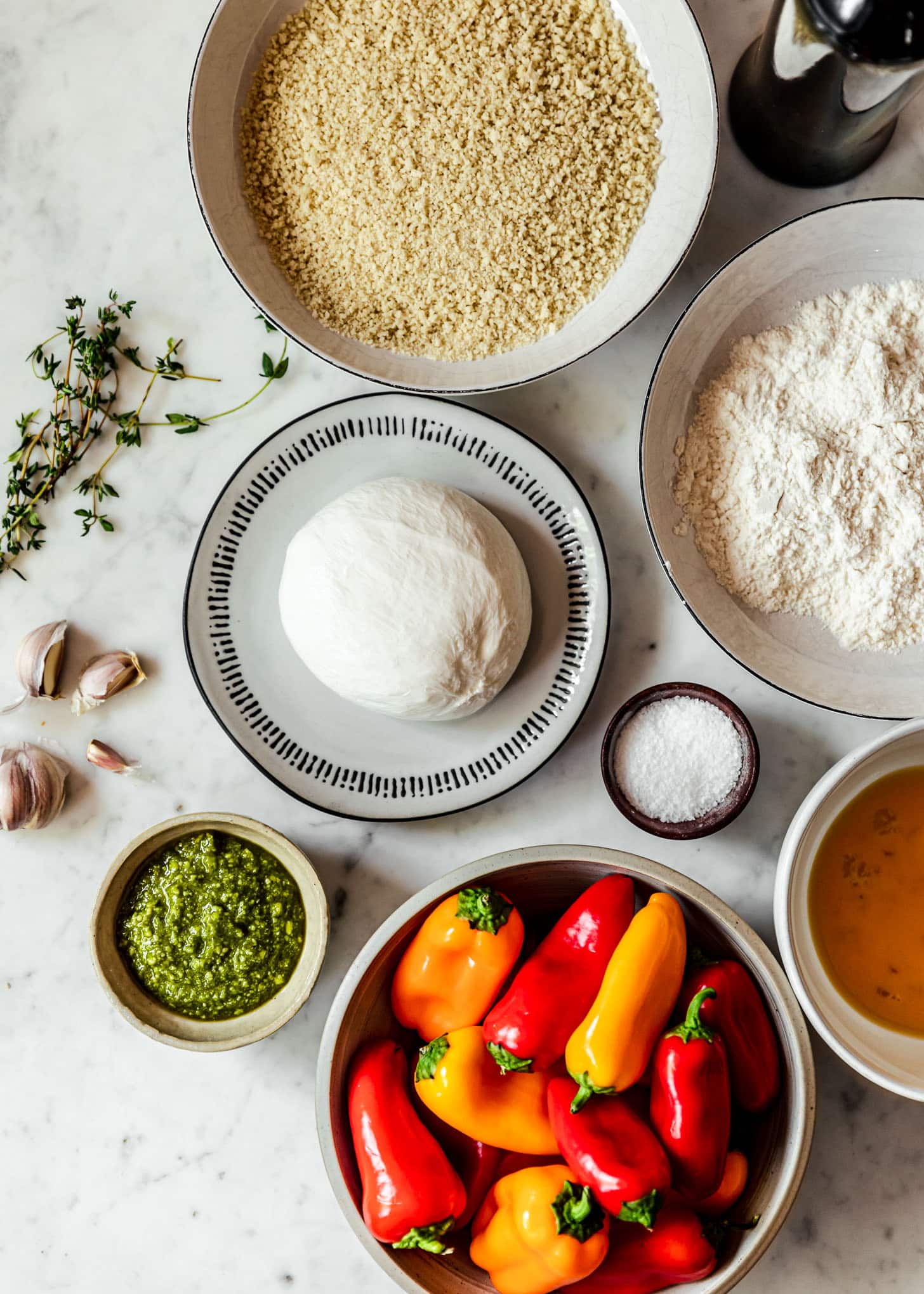 A plate of burrata on a white counter next to breadcrumbs, pesto, eggs, flour, and mini bell peppers.