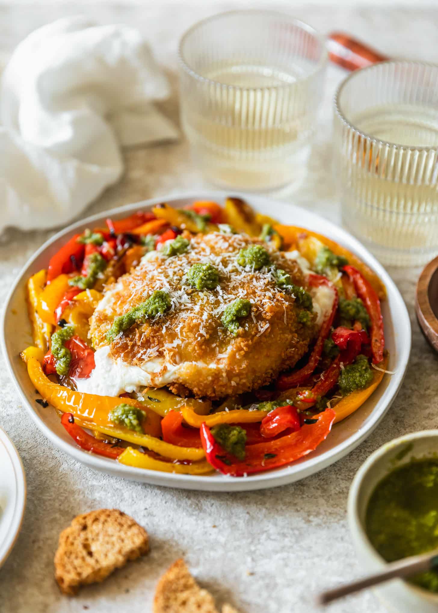 Fried burrata on a white plate with roasted peppers next to glasses of white wine, a white linen, and crackers with a beige background.