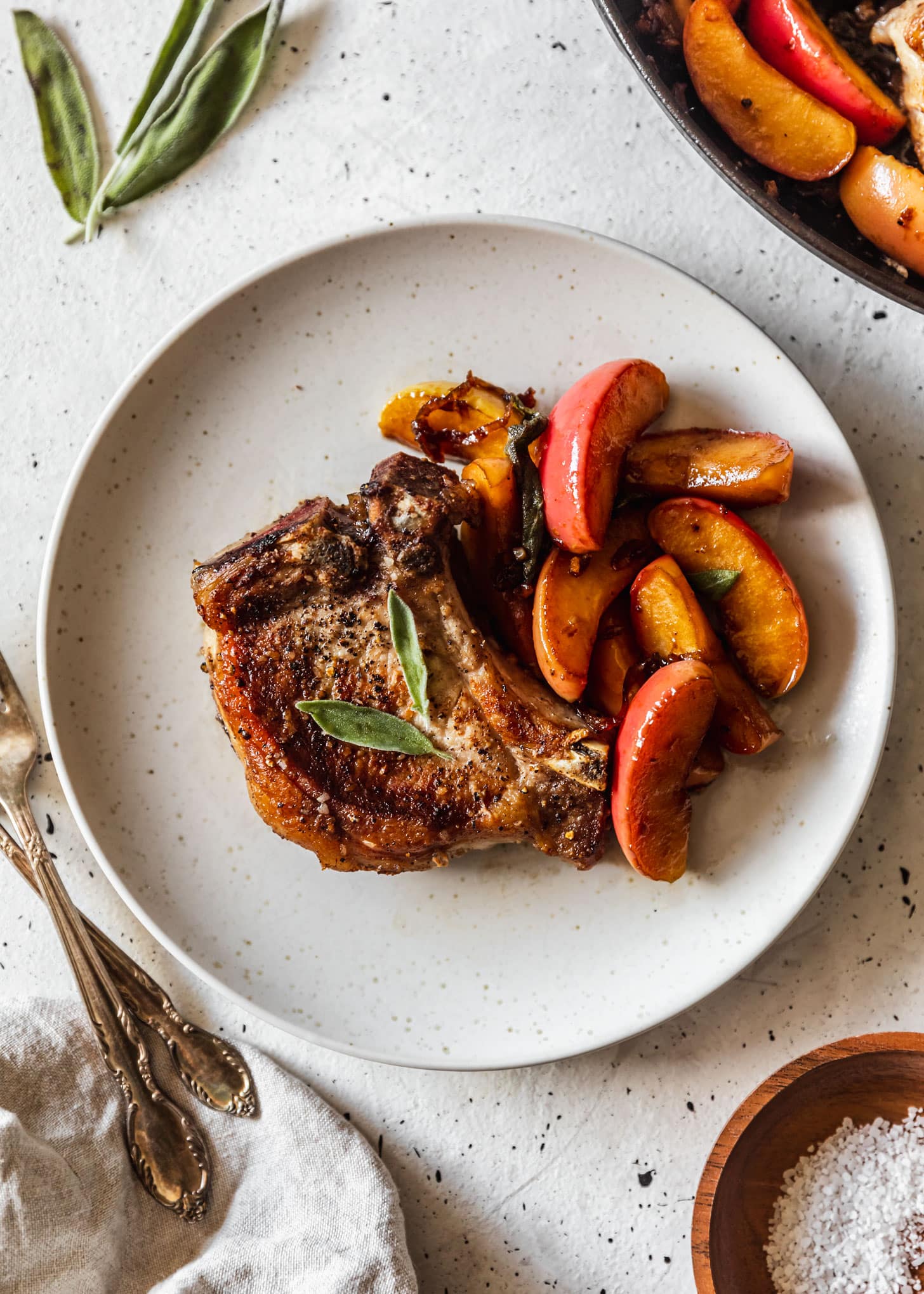 A pork chop with apples and sage on a white plate next to a pan of apple pork chops on a white speckled background.