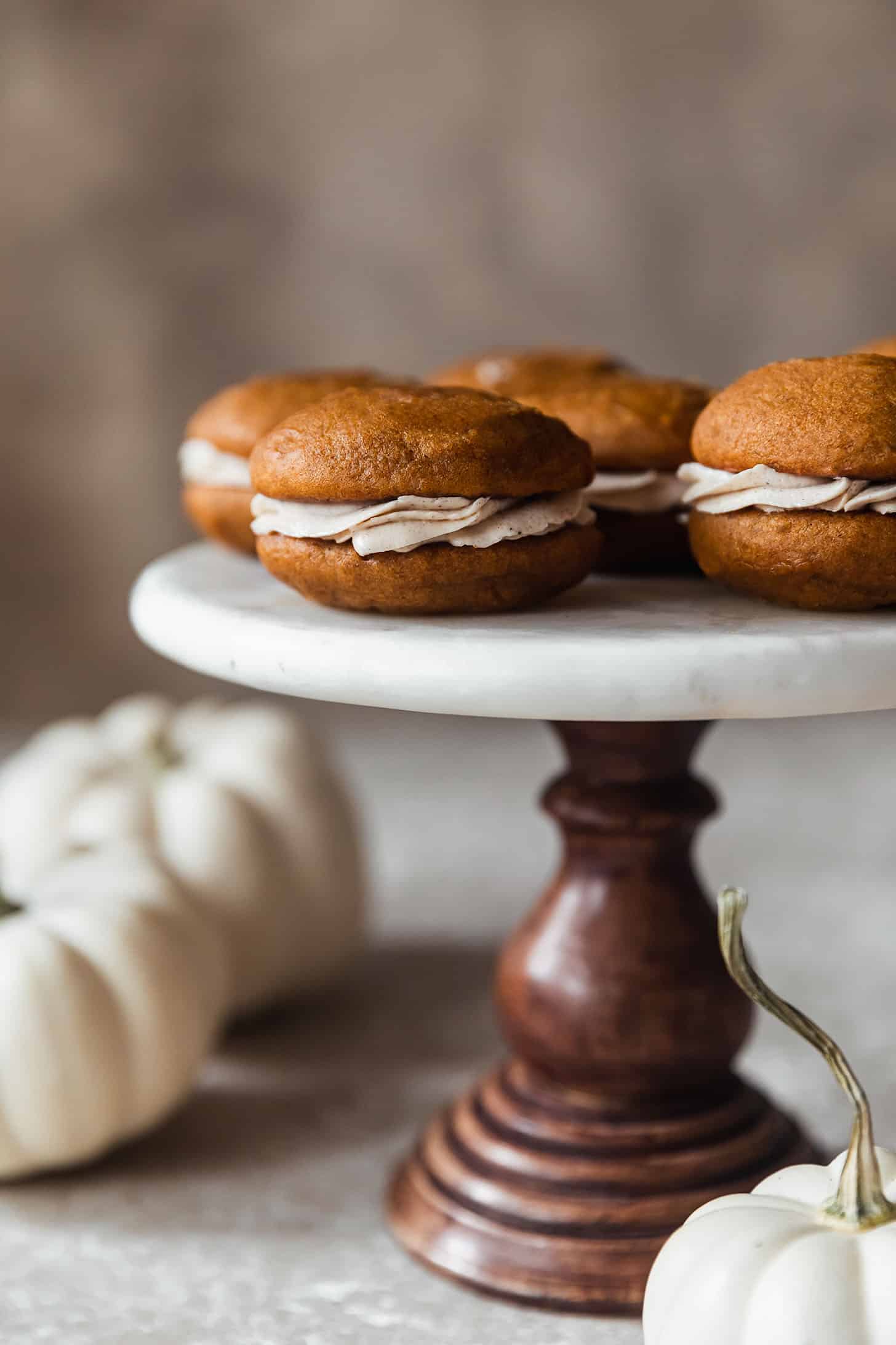 A closeup of pumpkin whoopie pies with chai buttercream on a marble and wood cake stand next to white pumpkins with a beige background.