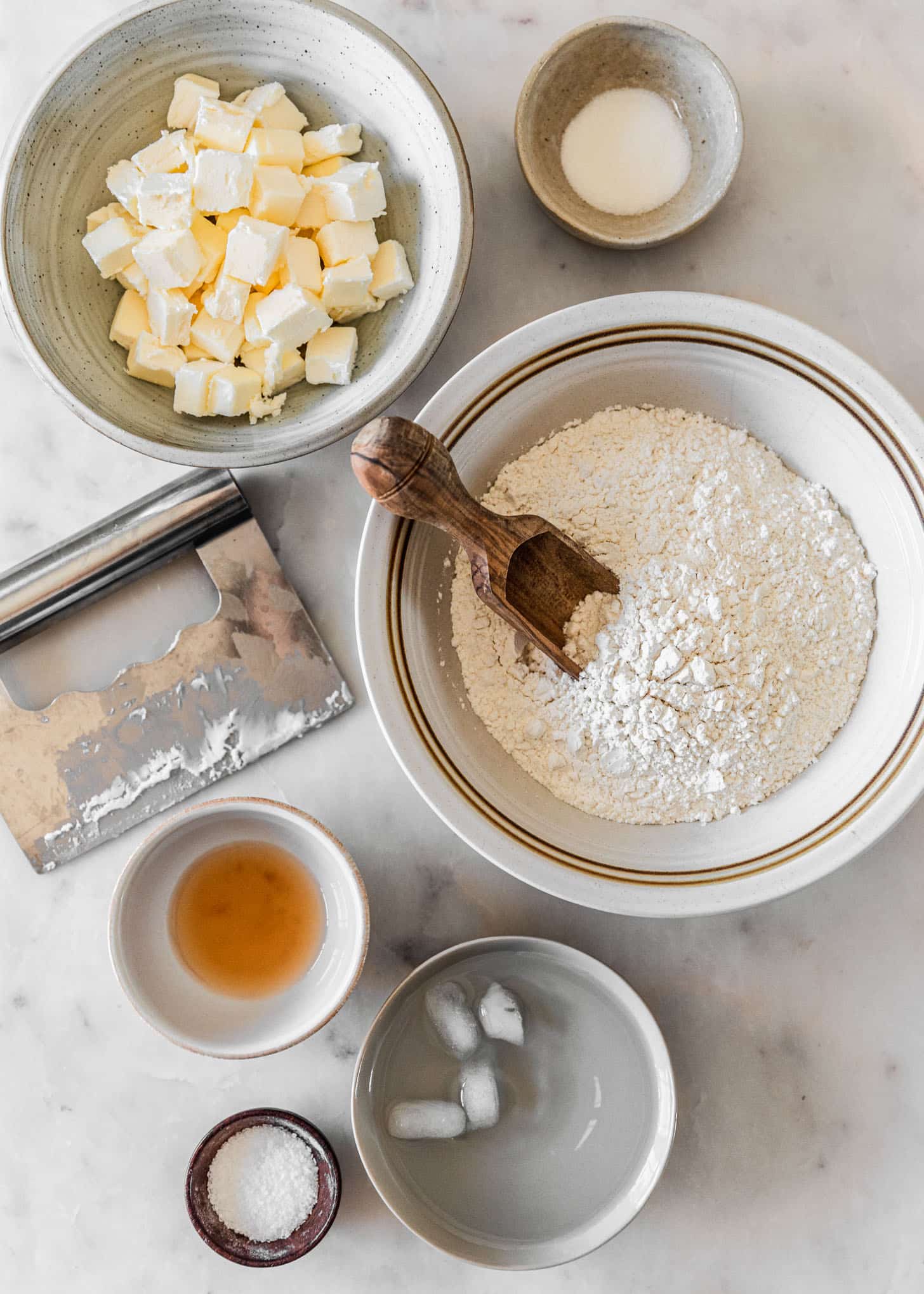 Ingredients for homemade pie dough in white and grey bowls on a white marble counter.