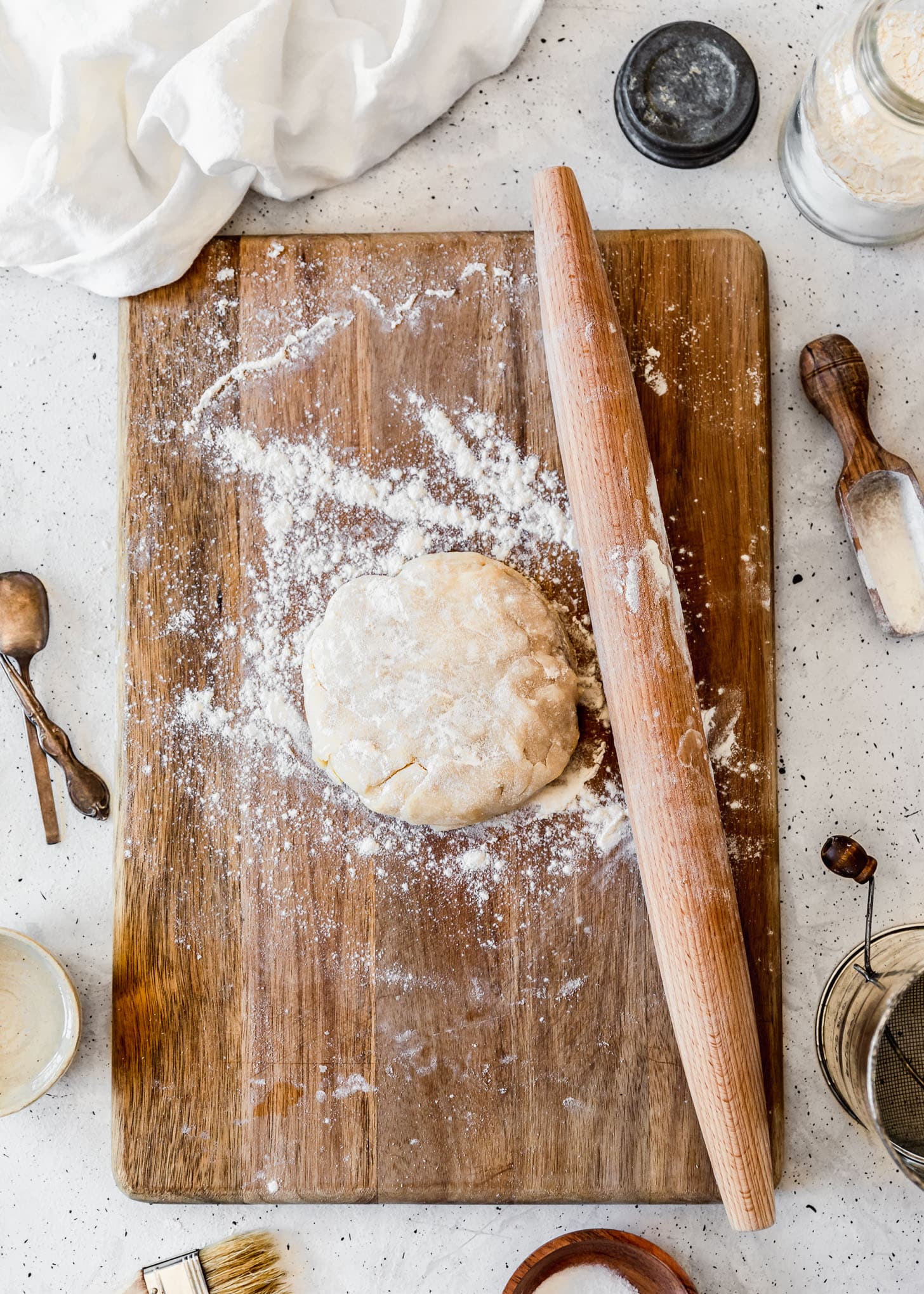 A disc of pie dough on a wood board surrounded by pie ingredients and tools with a white speckled background.
