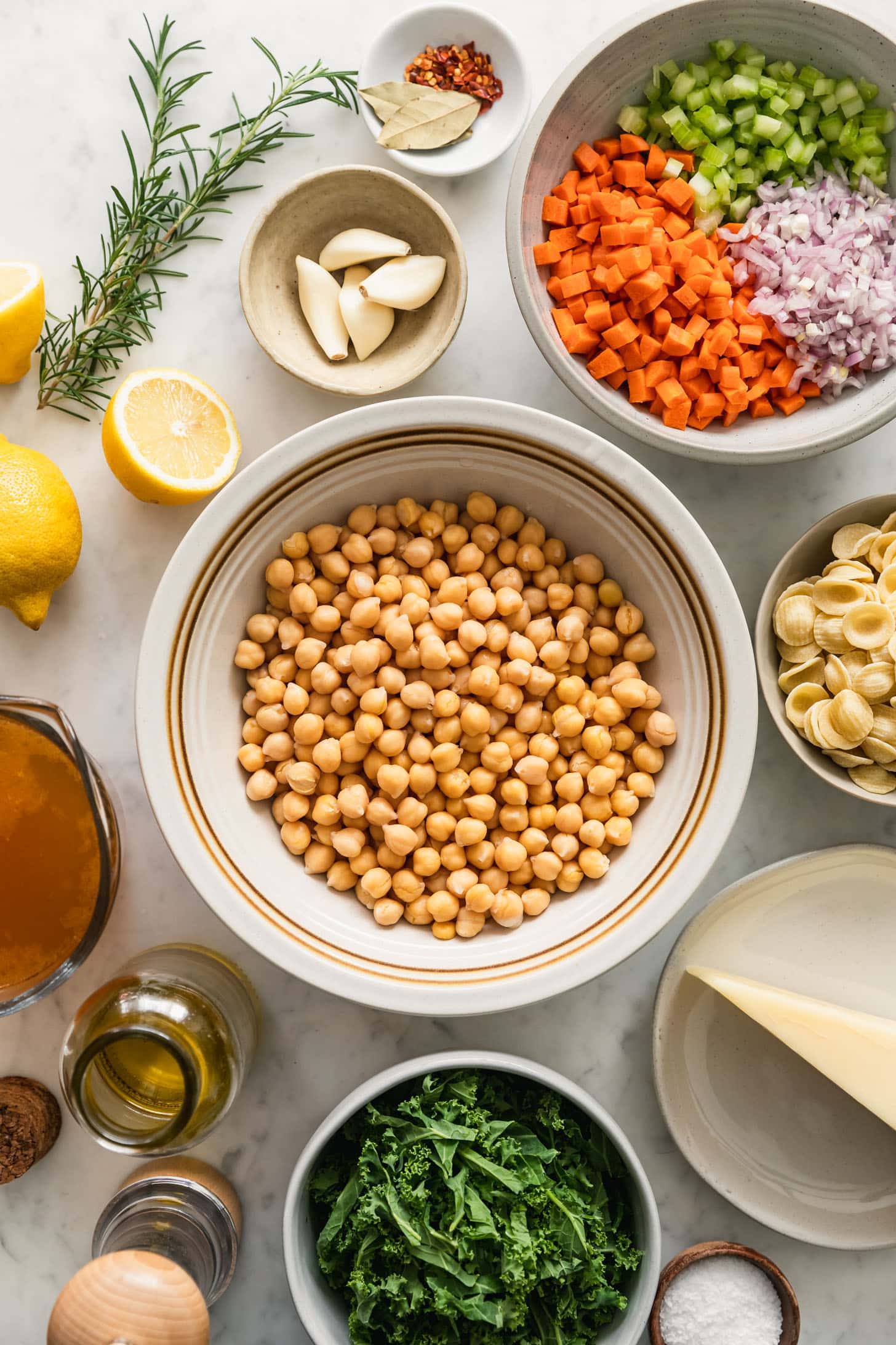 Chickpeas, veggies, pasta, Parmesan, and spices in white bowls next to lemons and rosemary on a white marble counter.