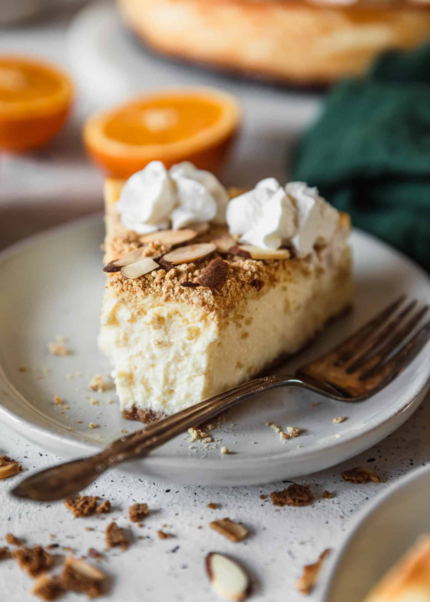 A closeup of mascarpone cheesecake with a bite taken out of it on a white plate with oranges and a green linen in the background.